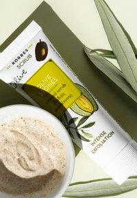 Korres Olive Stones skrubb rör i vitt med gröna detaljer, med en brun olivillustration. Åtföljd av en vit skål med exfolierande kräm.