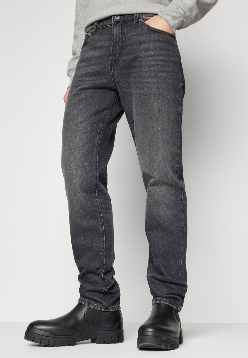 Osoba w wyblakłych czarnych slim-fit jeansach i czarnych masywnych botkach Chelsea, stojąca na tle jednolitej jasnej ściany.