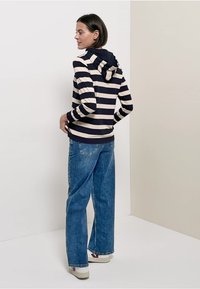 Navy and cream striped hoodie with a hood, paired with loose blue jeans and white sneakers with red accents. Rear view against a neutral background.