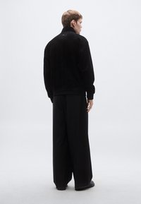 Μαύρο τζάκετ με χνουδωτή υφή και ψηλό γιακά, με μανσέτες με ρίγες και μαύρο φαρδύ παντελόνι. Ομαλή υφή, απλός σχεδιασμός και χωρίς ορατά λογότυπα.