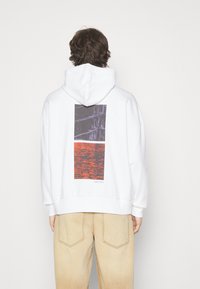 Felpa bianca con cappuccio caratterizzata da una stampa grafica sul retro con design rettangolari in texture viola scuro e rosso.