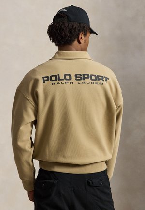 Mand iført beige Polo Sport Ralph Lauren trøje og sort kasket, set bagfra mod neutral baggrund.
