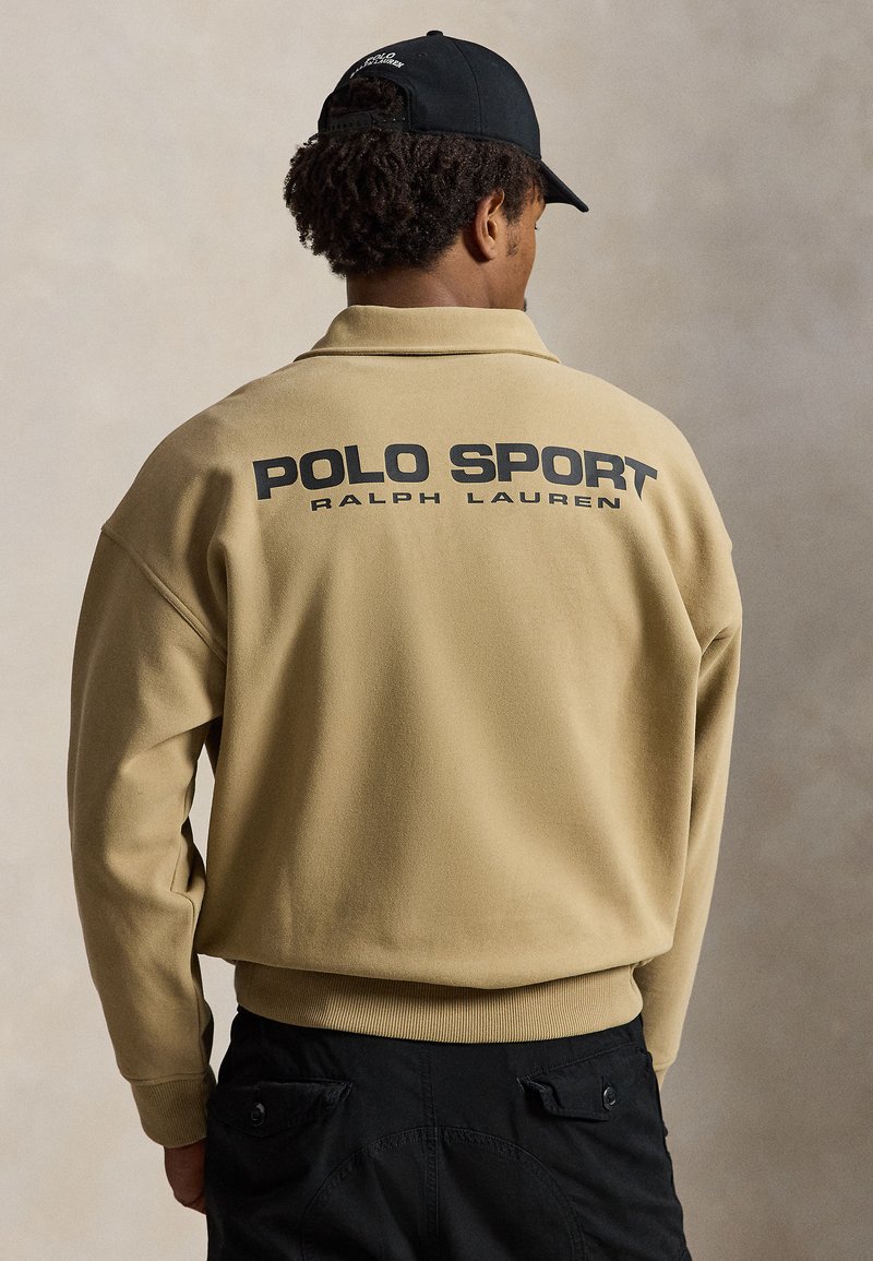 Άνδρας που φοράει μπεζ πουλόβερ Polo Sport Ralph Lauren και μαύρο καπέλο, εμφανίζεται από πίσω σε ουδέτερο φόντο.