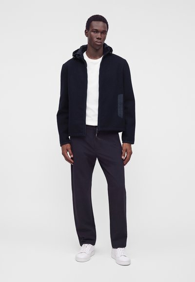 Homme portant une veste noire à capuche, un t-shirt blanc, un pantalon noir et des baskets blanches, debout devant un fond clair uni.