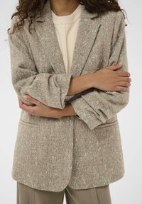 Bež blazer v keper vzoru, izdelan iz teksturirane tkanine, z pikastimi reverji, sprednjimi žepi in zavitimi rokavi. Mehka, prilagojena silhueta.