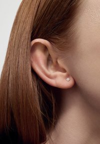 Małe, okrągłe kolczyki wkręty z przezroczystym kamieniem, osadzone w metalowej podstawie, noszone w przekłutym uchu, uzupełniające proste brązowe włosy.