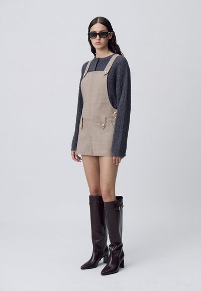 Vestido overall a cuadros beige con bolsillos laterales y botones dorados, combinado con un suéter de punto trenzado gris y botas marrones hasta la rodilla.
