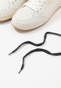 Hvide lædersneakers med perforeret tå, beige gummisål og flade sorte snørebånd med metalliske spidser. Enkel design med minimale accenter.