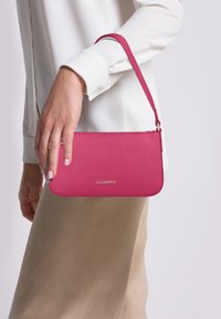 Bolso de mano de cuero rosa con un acabado texturizado, forma delgada y una correa desmontable. Presenta un logo dorado en el frente.