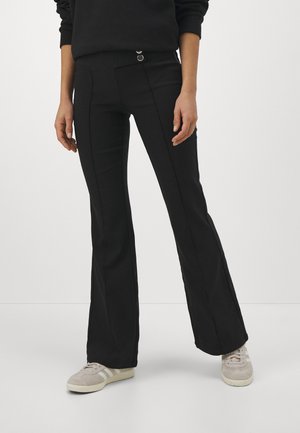 Persona con pantalones acampanados negros con costuras visibles, zapatillas claras y una camiseta negra de manga larga, de pie contra un fondo liso.