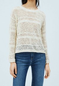 Femme portant un pull à manches longues en maille crème avec un motif ondulé ajouré, associé à un jean en denim bleu.