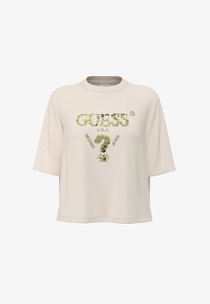 T-shirt beige a maniche corte con la scritta "GUESS U.S.A." e un disegno di un punto interrogativo con paillettes, inclusi i dettagli testuali "Washed" e "Jeans".