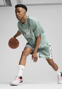 Mintgrünes Sport-T-Shirt und Shorts mit weißem Puma-Logo. Weiße Socken und rot akzentuierte schwarze Basketballschuhe. Hält einen Basketball.