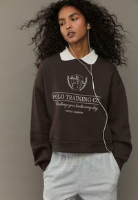Felpa marrone con colletto bianco, caratterizzata da un logo e testo ricamati. Indossata con pantaloni della tuta grigi e auricolari bianchi.