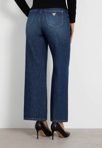 Jeans de mezclilla oscuros de pierna ancha con dobladillo ajustado, que cuentan con dos bolsillos traseros y un pequeño detalle de logo. Llevados con tacones altos negros.