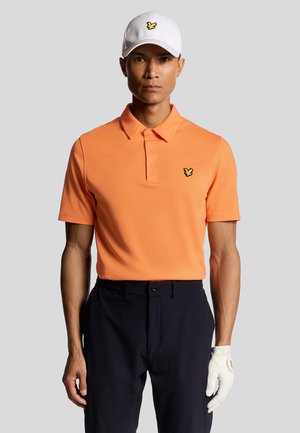Orange kortærmet polo-shirt med krave og gult sommerfuglelogo, parret med mørke bukser og en hvid kasket. En hånd i handske er synlig.