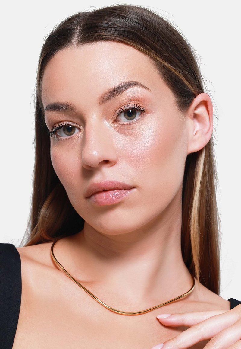 Gullmetall choker halskjede med et glatt, buet design, båret mot mørkt klær, som fremhever nakkeområdet. Subtil glans og elegant form.