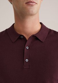 Férfi sötét bordó pólót visel, három fekete gombbal, látható nyakkal és alsó arccal egy semleges háttér előtt.