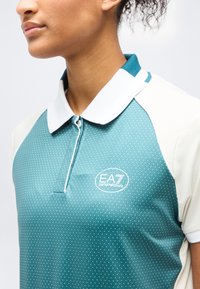 Κοντινό πλάνο ατόμου που φοράει μπλε-πράσινο και λευκό πουά πόλο μπλουζάκι EA7 Emporio Armani με λευκό γιακά και κοντά μανίκια.