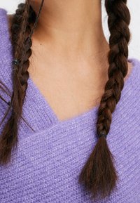 Maglione lavorato a maglia lavanda con design a V, caratterizzato da una consistenza morbida e una vestibilità ampia, abbinato a lunghe trecce marroni fissate con elastici.