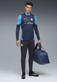 Echipament de antrenament de fotbal Puma, cu un top pe mânecă lungă, modelat în nuanțe de albastru închis și colanți negri, purtând o geantă sport bleumarin în mână și ghete de fotbal.
