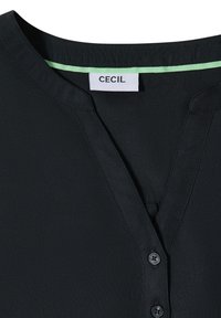 Blouse noire col en V avec une étiquette de marque indiquant "CECIL" à l'intérieur de l'encolure et deux boutons noirs visibles sous le col.