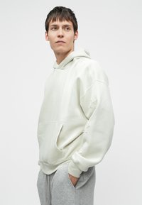 Lys grå oversized hettegenser med kengurulomme, ribbede mansjetter og diskret stjernebroderi på ermene. Mykt, teksturert stoff.