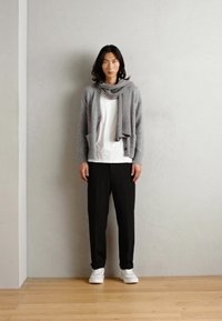 Cardigan gri peste un tricou alb, pantaloni negri și adidași albi. Păr lung și negru, stând lângă un zid gri, cu podea din lemn.