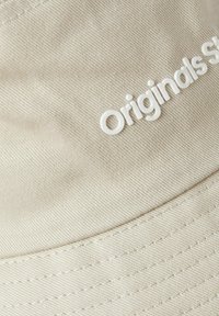 Beige cap made of textured fabric with a curved brim. Features raised white lettering stating "Originals." Stitching along the brim is visible.