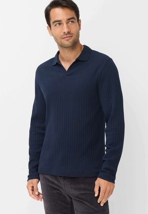 Uomo che indossa una polo a costine blu scuro a maniche lunghe e pantaloni scuri, in piedi davanti a uno sfondo chiaro e uniforme.