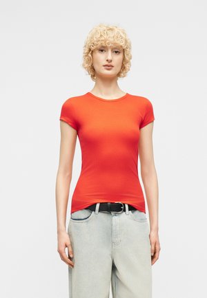 Γυναίκα με κοντά σγουρά ξανθά μαλλιά που φοράει εφαρμοστό φωτεινό πορτοκαλί κοντομάνικο πουκάμισο και ανοιχτόχρωμο τζιν με μαύρη ζώνη.