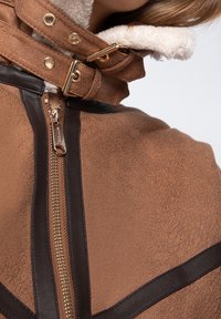 Veste en faux suède marron avec un épais col en fourrure blanche. Présente une fermeture éclair à l'avant et des accents en métal doré. Détails en cuir brun foncé.