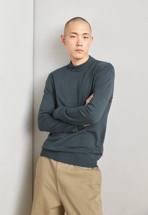 Maglione in maglia di un verde scuro, con colletto alto, maniche lunghe e polsini a coste, abbinato a pantaloni beige. Texture liscia e design aderente.