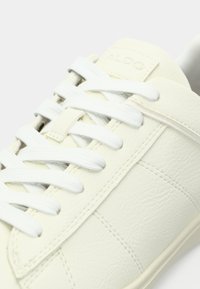 Baskets blanches en cuir végan avec surface texturée, lacets blancs et un logo discret sur la languette. Design minimaliste et détails de couture.