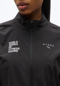 Κοντινό πλάνο ενός ατόμου που φοράει μαύρο τζάκετ με φερμουάρ, με τα λογότυπα «World Series of Fitness Racing» και «HYROX» της Puma στο στήθος.
