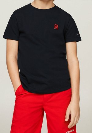 Kind trägt ein schwarzes T-Shirt mit rotem Logo auf der Brust und rote Shorts, eine Hand in der Tasche, steht vor einem neutralen Hintergrund.