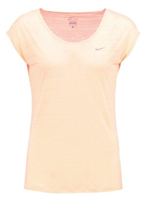 Kobieta idąca, ubrana w brzoskwiniową bluzkę z krótkim rękawem, czarne sportowe spodenki i czarne buty do biegania Nike na białym tle.