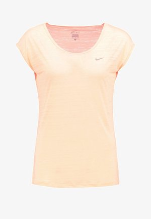 Kobieta idąca, ubrana w brzoskwiniową bluzkę z krótkim rękawem, czarne sportowe spodenki i czarne buty do biegania Nike na białym tle.