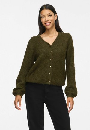 Donna che indossa un cardigan peloso verde scuro con bottoni di perla e pantaloni neri, sorridente con le mani rilassate lungo i fianchi.