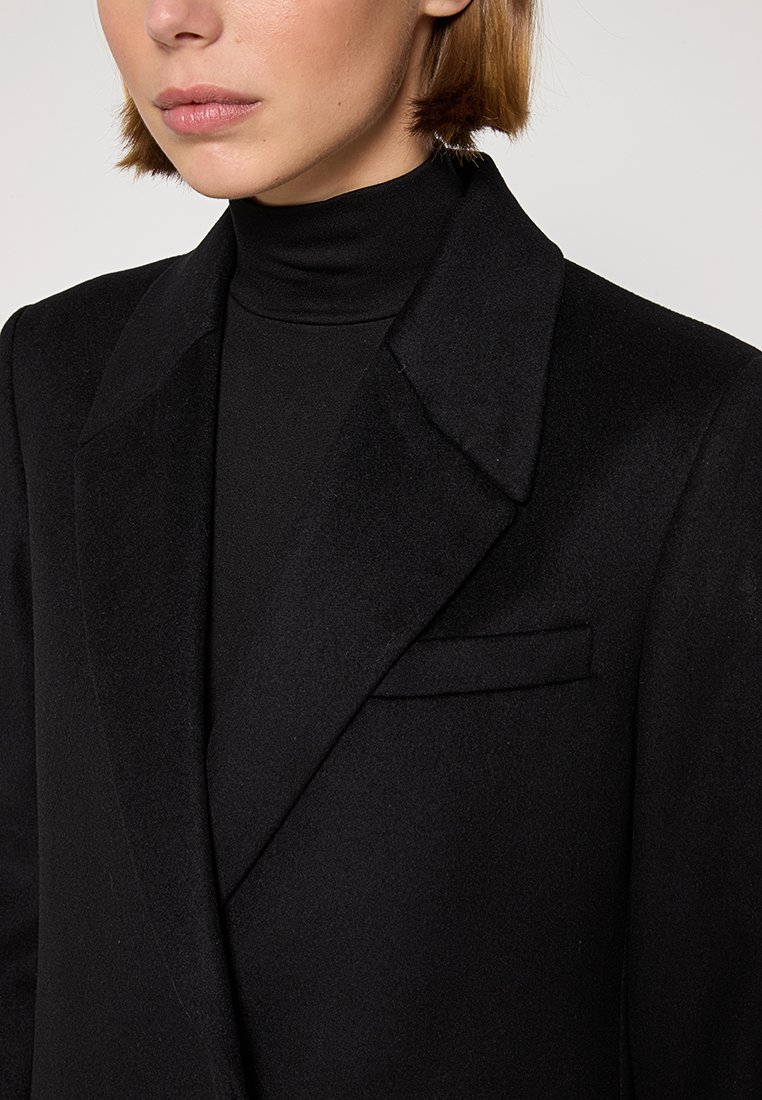 Blazer noir en mélange de laine avec des revers crantés, une poche plaquée unique et une texture douce et lisse. Porté par-dessus un col roulé noir.