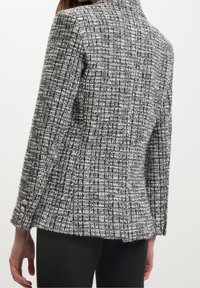 Blazer in tweed nero e bianco a trama, con silhouette aderente, colletto a revers e tre bottoni decorativi sui polsini.