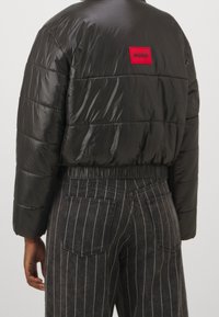 Vadderad svart jacka med kort design, elastisk fåll och en röd HUGO-logotyp på baksidan. Kombinerad med grå randiga jeans.