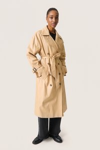 Beige trenchcoat med strop i taljen, stor krave og dobbeltknaplukning, med mørke knapper og justerbare manchetter. Bæres over sorte bukser.