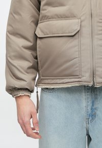 Dunjakke i lys beige med tekstureret finish, med lynlåslukning, sidelommer og elastiske manchetter. Båret over lyseblå jeans.