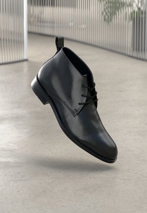 Schwarze Lederstiefelette mit glatter Oberfläche, runder Zehenpartie und schnürbarem Vorderteil. Mit einem kleinen Absatz und einem Zugband an der Rückseite für einfaches Anziehen.