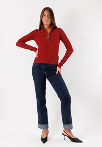 Maglione rosso con zip e texture a costine, dotato di una cerniera a un quarto di lunghezza. Abbinato a jeans blu scuro con risvolto e tacchi a punta neri.