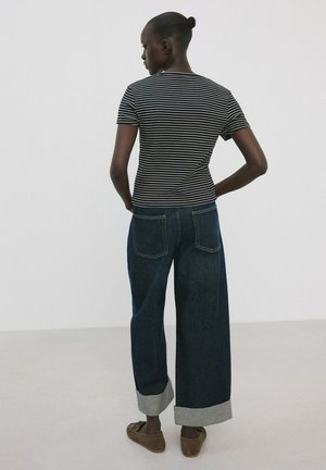 Personne debout de dos portant un t-shirt rayé noir et blanc, un jean foncé large avec des revers et des chaussures marron sans lacets.