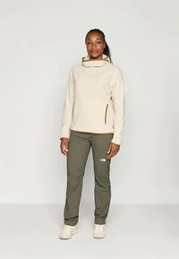 Pullover di pile beige con colletto alto e zip laterale, abbinato a pantaloni verde oliva con logo. Le scarpe in suede completano l'outfit.