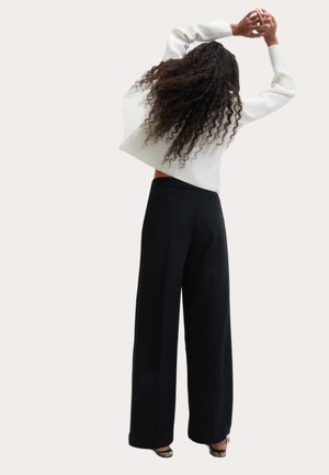 Vrouw met lang krullend haar die haar armen boven haar hoofd uitstrekt, gekleed in een witte top met lange mouwen, zwarte wijde broekspijpen en hakken met slangenprint.