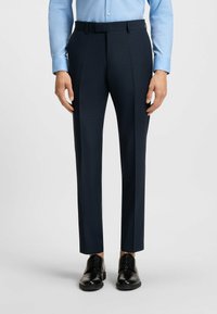 Marineblaue Anzughose aus strukturiertem Stoff, mit taillierter Passform, Gürtelschlaufen und einem Falten in der Mitte, kombiniert mit schwarzen Anzugschuhen.